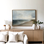 A minimalist living room features a light wood sideboard with white and beige vases, a lamp, and the Shoreline Calm Canvas Art displayed above. A white sofa with neutral pillows completes the serene coastal decor.