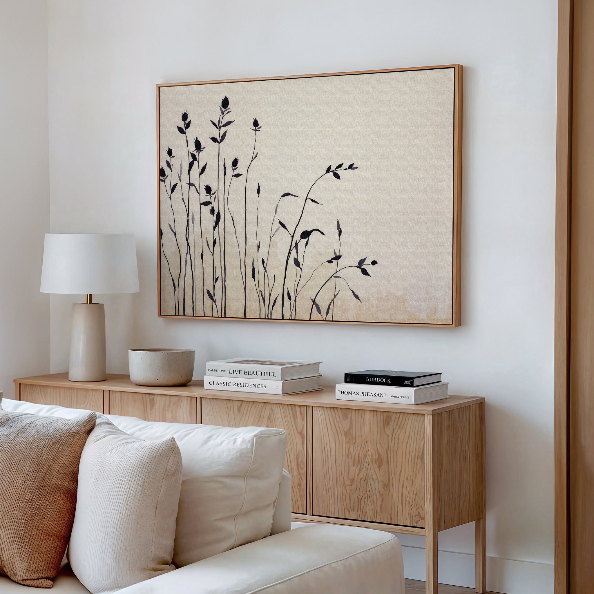 A modern living room with a light wood sideboard, stacked books, a beige bowl, and a white lamp is accented by Nordic decor featuring the Silhouette Meadow Canvas Art - Horizontal above the sideboard—black plant silhouettes on a neutral background.
