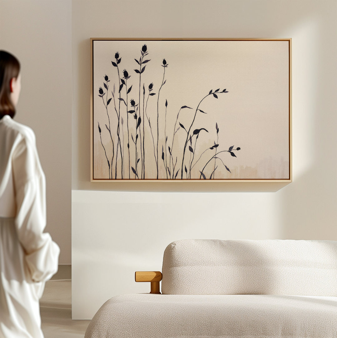 A person in a light outfit stands in a minimalist room, admiring the Silhouette Meadow Canvas Art - Horizontal, featuring tall black floral silhouettes on beige, displayed above a light-colored sofa.