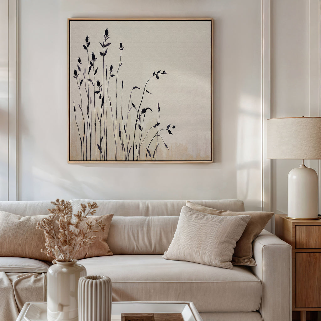 A modern living room features a beige sofa with neutral cushions, a light wood side table, and a white lamp. Above, the Silhouette Meadow Canvas Art - Square adds Nordic flair, while dried flowers in a vase accent the coffee table.
