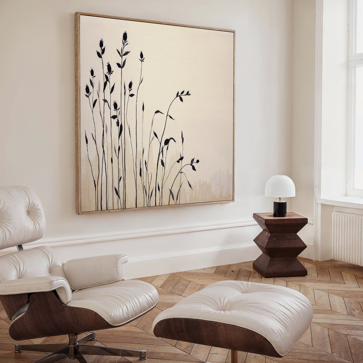 A modern living room features a white leather lounge chair and ottoman, a dark wood side table with a white lamp, and the Silhouette Meadow Canvas Art - Square—an elegant complement to minimalist Nordic decor.