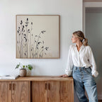 A woman in a white blouse and jeans leans on a wooden cabinet, admiring the Silhouette Meadow Canvas Art - Square. A small potted plant and book nearby enhance the calm Nordic decor of the modern room.