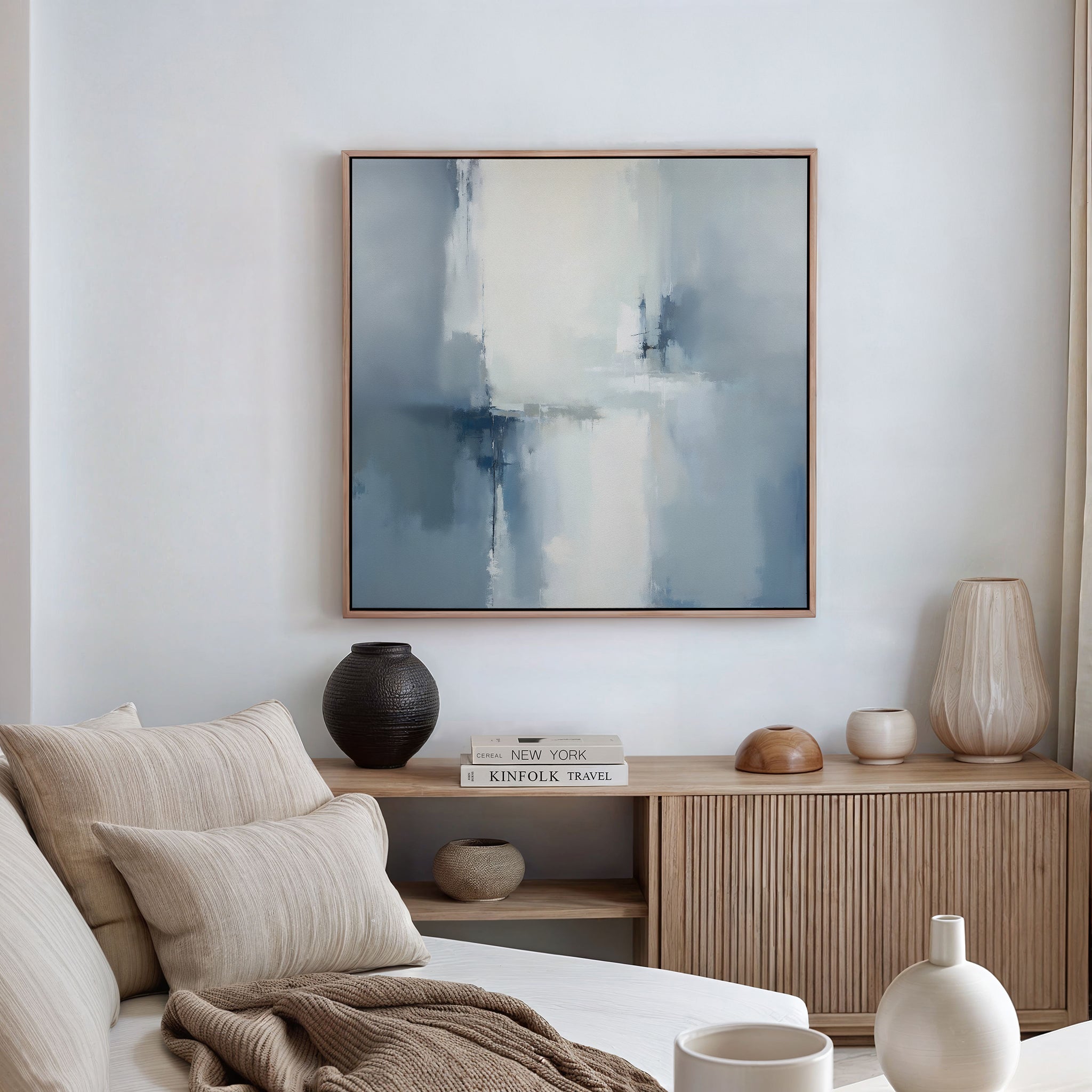 A modern living room featuring a beige couch, striped pillows, wooden sideboard, decorative vases, stacked books, and the Silver Cross Formation Canvas Art - Square on the wall for a Nordic-inspired touch.
