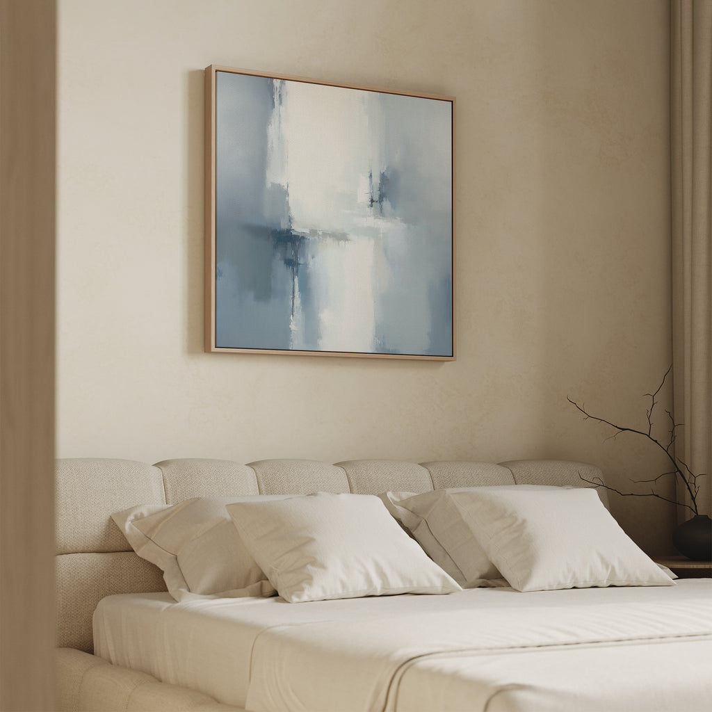 A minimalist bedroom features a cream upholstered bed with white pillows. Above the bed, the Silver Cross Formation Canvas Art - Square in soft blue and white tones creates a serene focal point. A dark vase with bare branches sits on the nightstand.