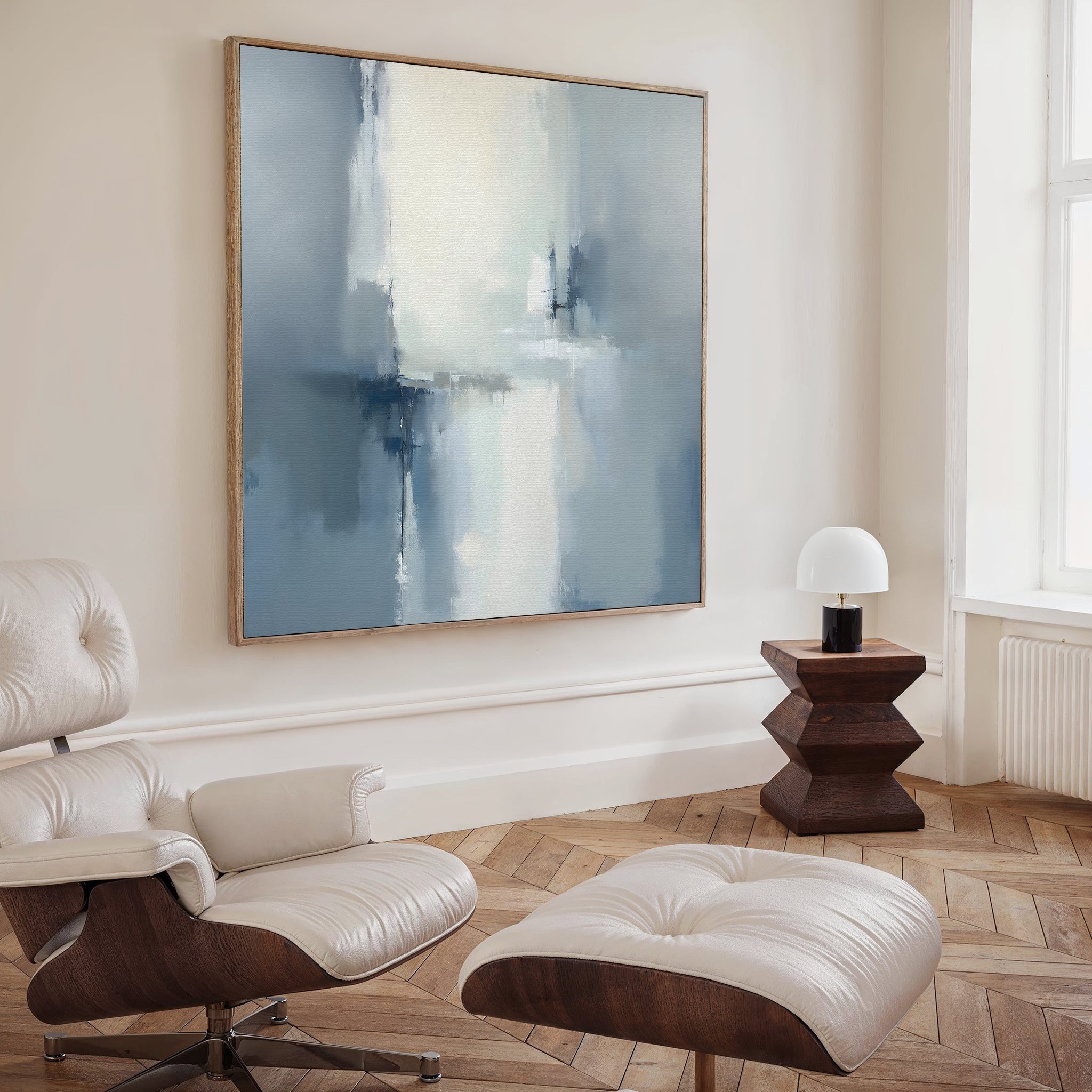 A modern living room featuring a white leather lounge chair and ottoman, a geometric wooden side table with a small white lamp, and the Silver Cross Formation Canvas Art - Square in blue and white on the wall.