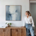 A woman in a white blouse and jeans leans on a wooden cabinet, admiring the Silver Cross Formation Canvas Art - Square. A small plant and a book rest on the cabinet beneath this modern abstract print, ideal for contemporary living rooms.