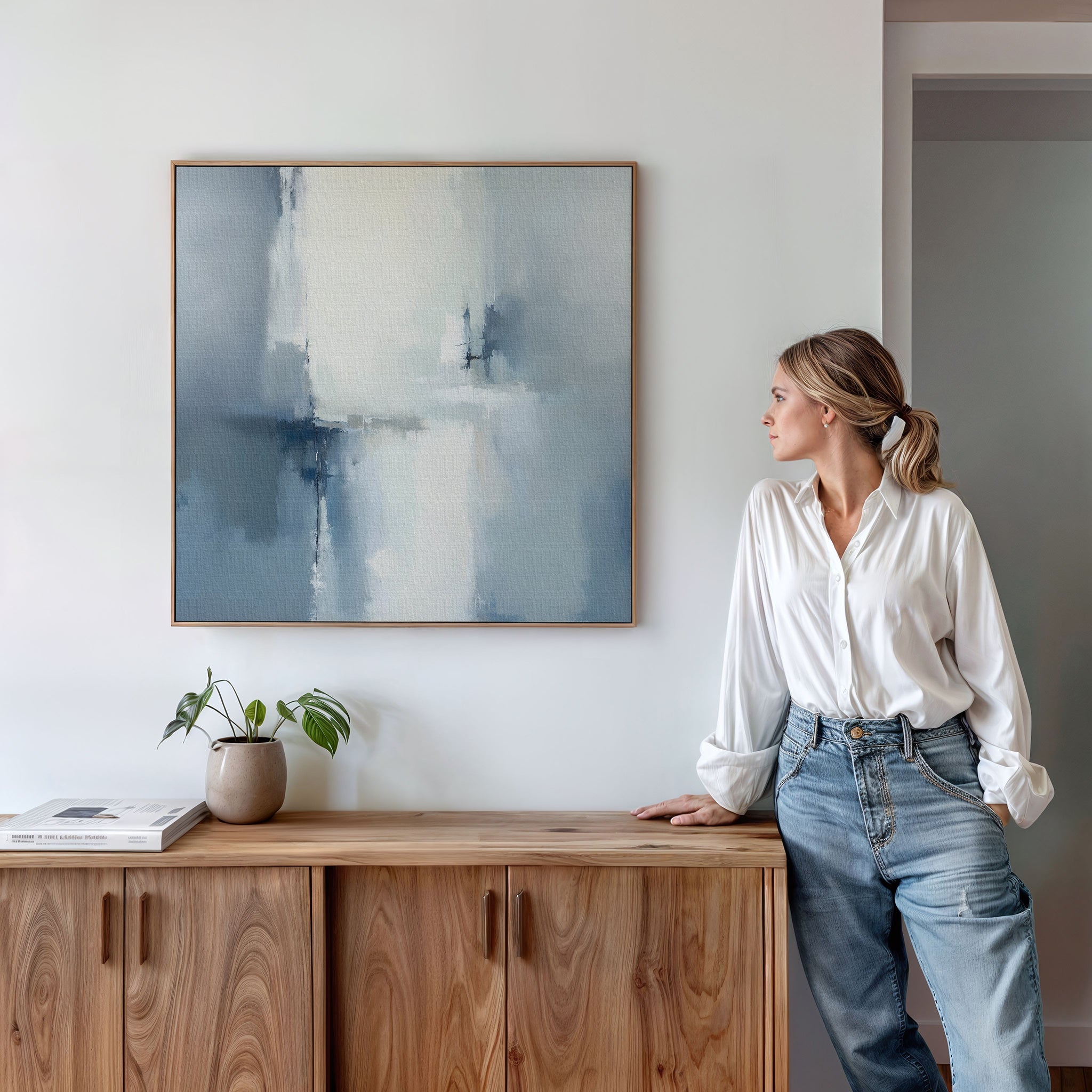 A woman in a white blouse and jeans leans on a wooden cabinet, admiring the Silver Cross Formation Canvas Art - Square. A small plant and a book rest on the cabinet beneath this modern abstract print, ideal for contemporary living rooms.