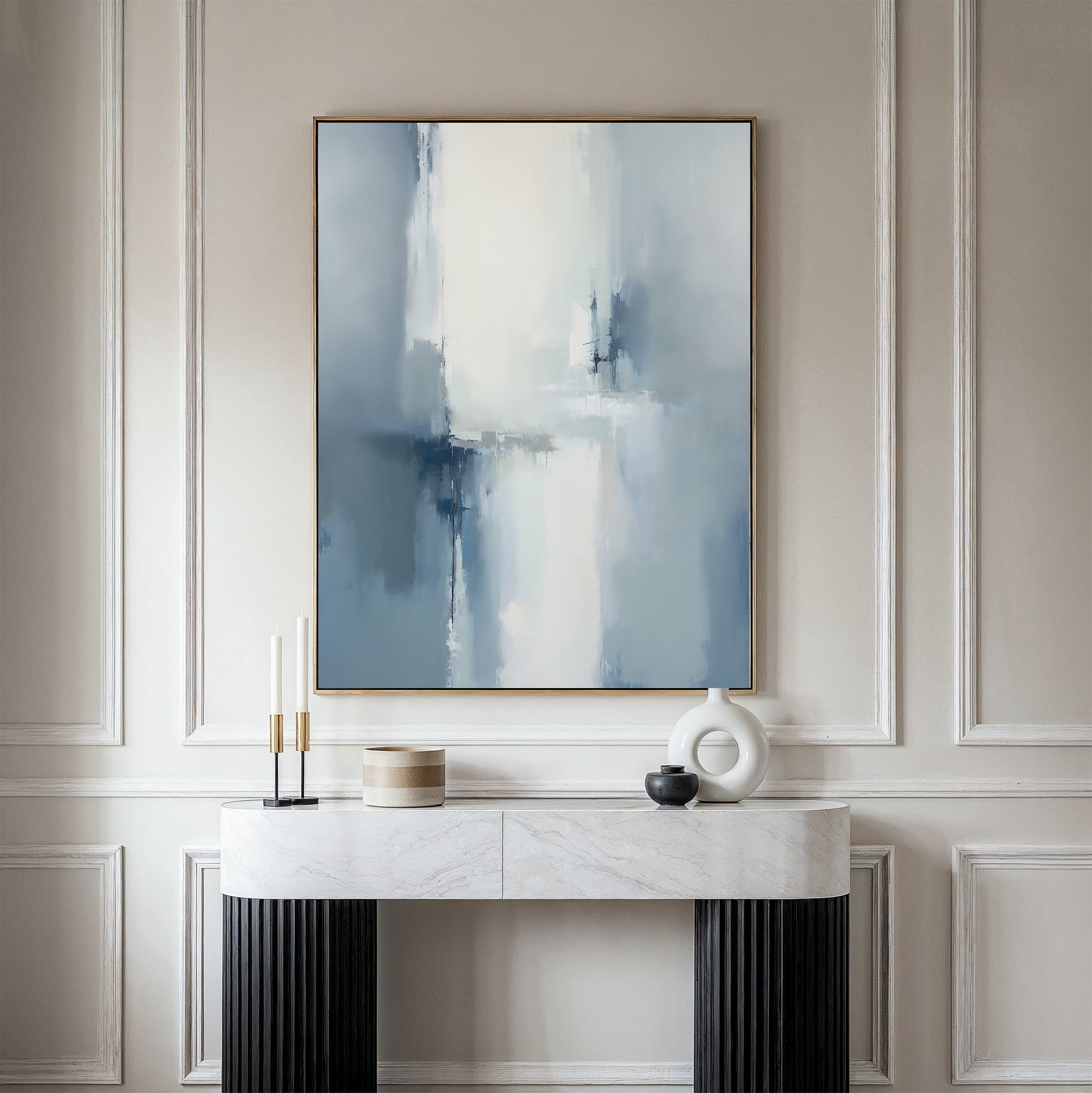 A modern entryway table with decor sits against a light paneled wall, topped by the Silver Cross Formation Canvas Art - Vertical, featuring atmospheric abstract tones of blue, white, and gray in a gold frame.
