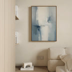 A modern living room corner featuring a beige sofa, a wooden bookshelf with books, and the "Silver Cross Formation Canvas Art - Vertical" in a wooden frame displayed on a light-colored wall.