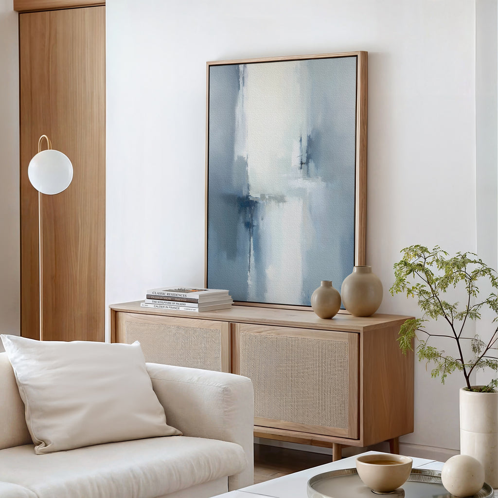 A modern living room with a light wood sideboard, Silver Cross Formation Canvas Art - Vertical, beige vases, stacked books, a leafy plant, and a white sofa with a pillow. A globe-shaped floor lamp stands beside the wooden door.