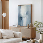 A modern living room with a light wood sideboard, Silver Cross Formation Canvas Art - Vertical, beige vases, stacked books, a leafy plant, and a white sofa with a pillow. A globe-shaped floor lamp stands beside the wooden door.