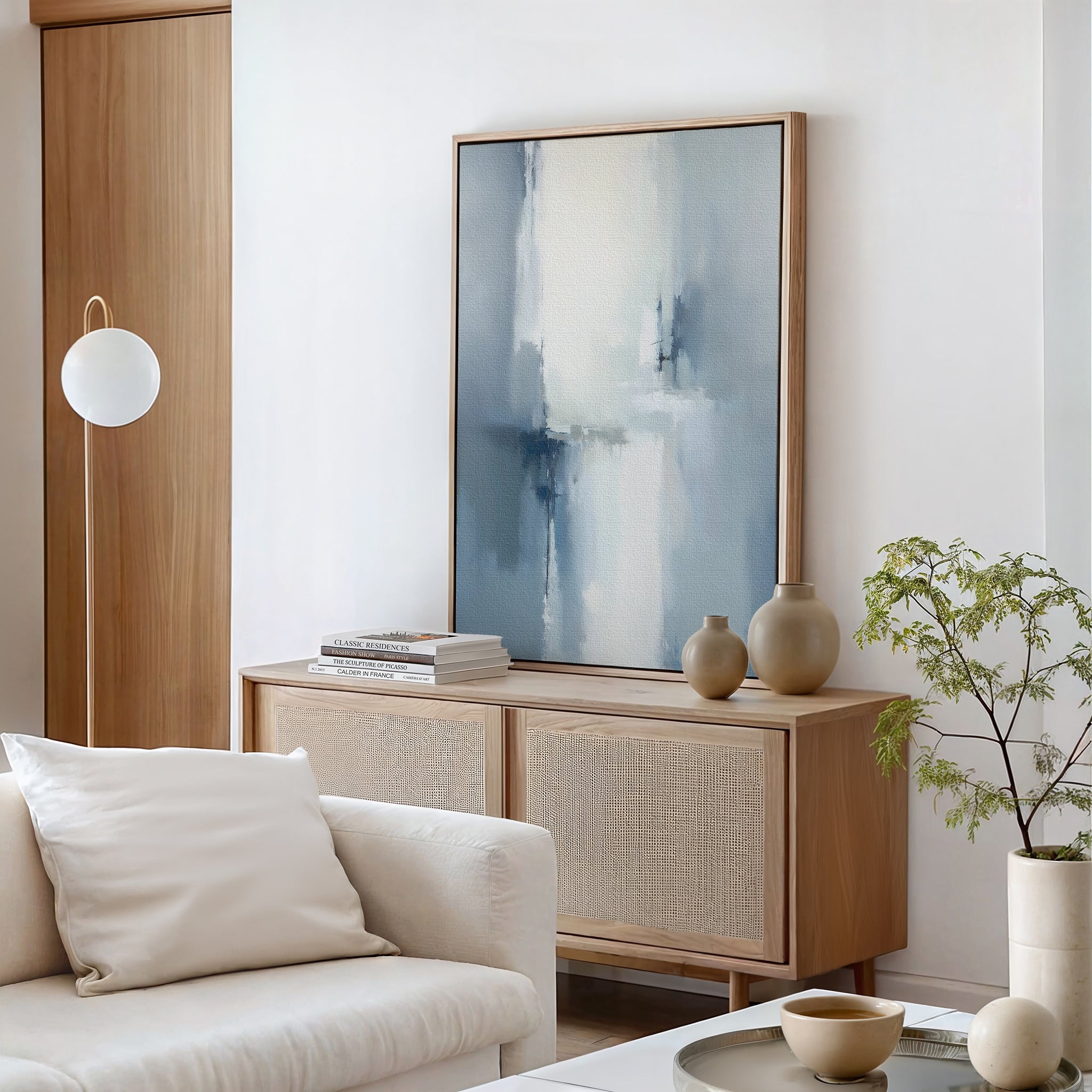 A modern living room with a light wood sideboard, Silver Cross Formation Canvas Art - Vertical, beige vases, stacked books, a leafy plant, and a white sofa with a pillow. A globe-shaped floor lamp stands beside the wooden door.