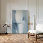A minimalist living space with herringbone wood floors, a white cushioned chair, a round side table with candles and books, and the Silver Cross Formation Canvas Art - Vertical leaning against a white paneled wall—ideal for modern living rooms.