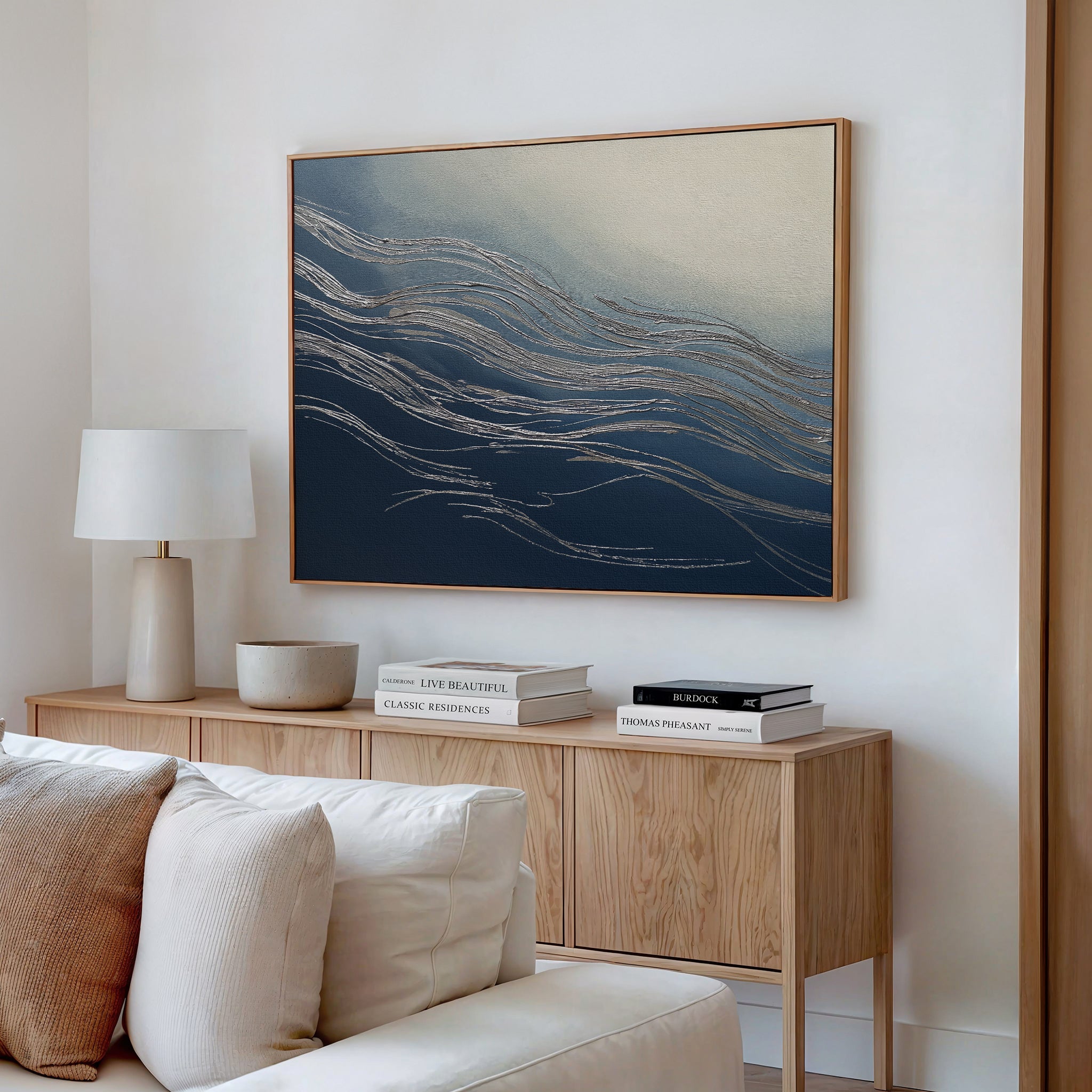 A modern living room features a beige sofa, a light wooden sideboard with books and décor, a white lamp, and the Silver Current Canvas Art - Horizontal with blue, gold, and cream wave-like patterns for contemporary sophistication.