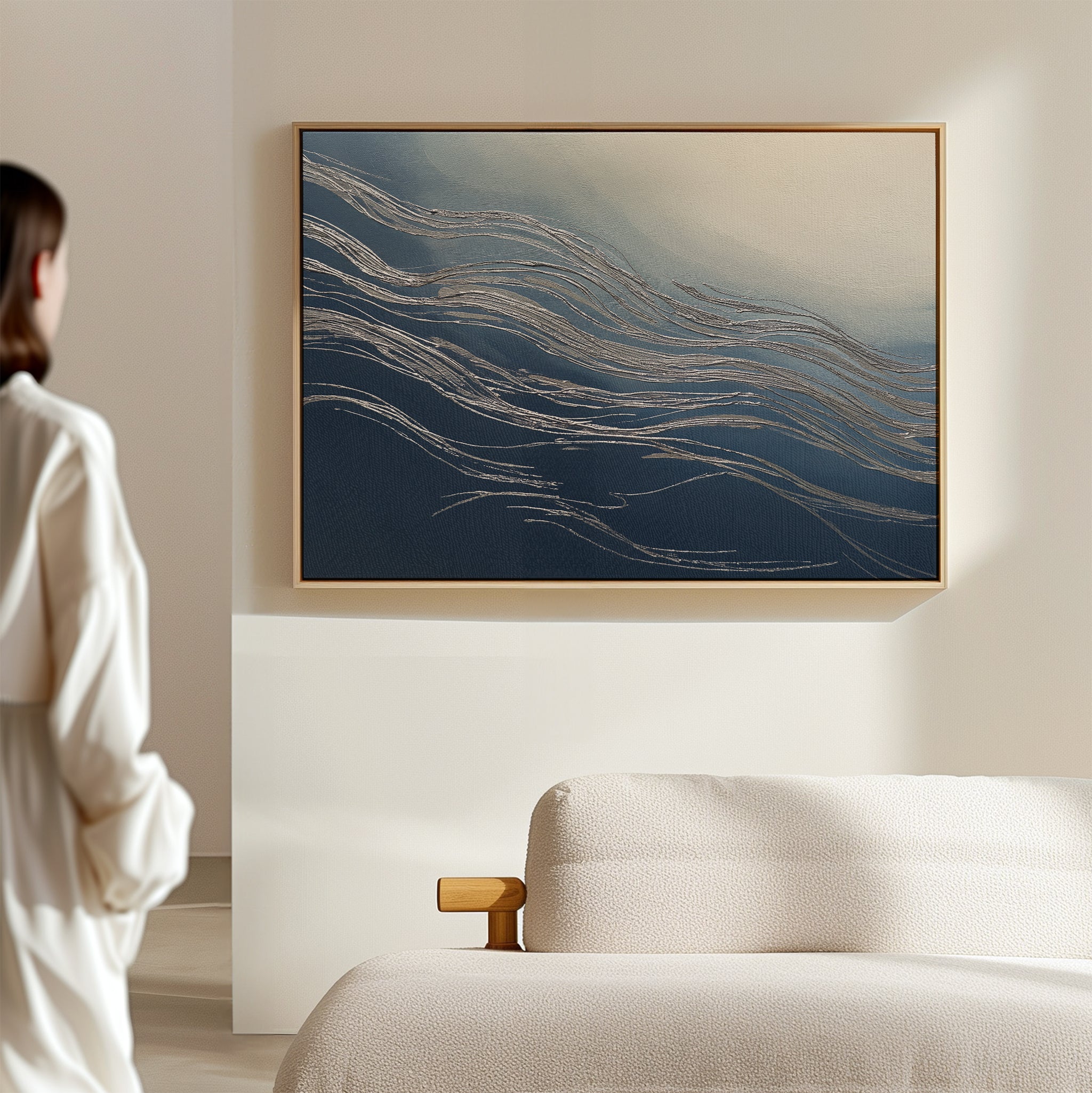 A person in a light outfit stands in a minimal, modern living room, admiring the Silver Current Canvas Art - Horizontal with flowing blue and beige lines above a beige sofa—a touch of contemporary sophistication.