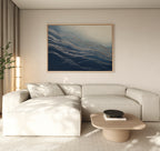 A modern living room with a light beige sectional sofa, a low wooden coffee table, a tall plant, and the Silver Current Canvas Art - Horizontal above the sofa adds contemporary sophistication with its blue and white abstract design.