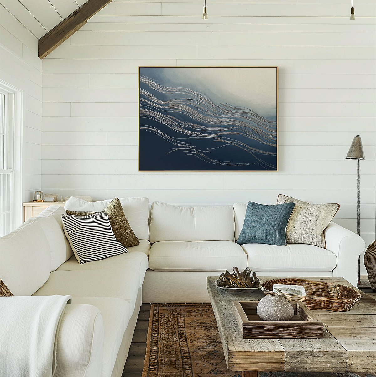 A cozy living room featuring a white sectional sofa, assorted pillows, a wooden coffee table with decor, and the Silver Current Canvas Art - Horizontal with blue and gold tones on a white shiplap wall. Natural light adds contemporary sophistication.