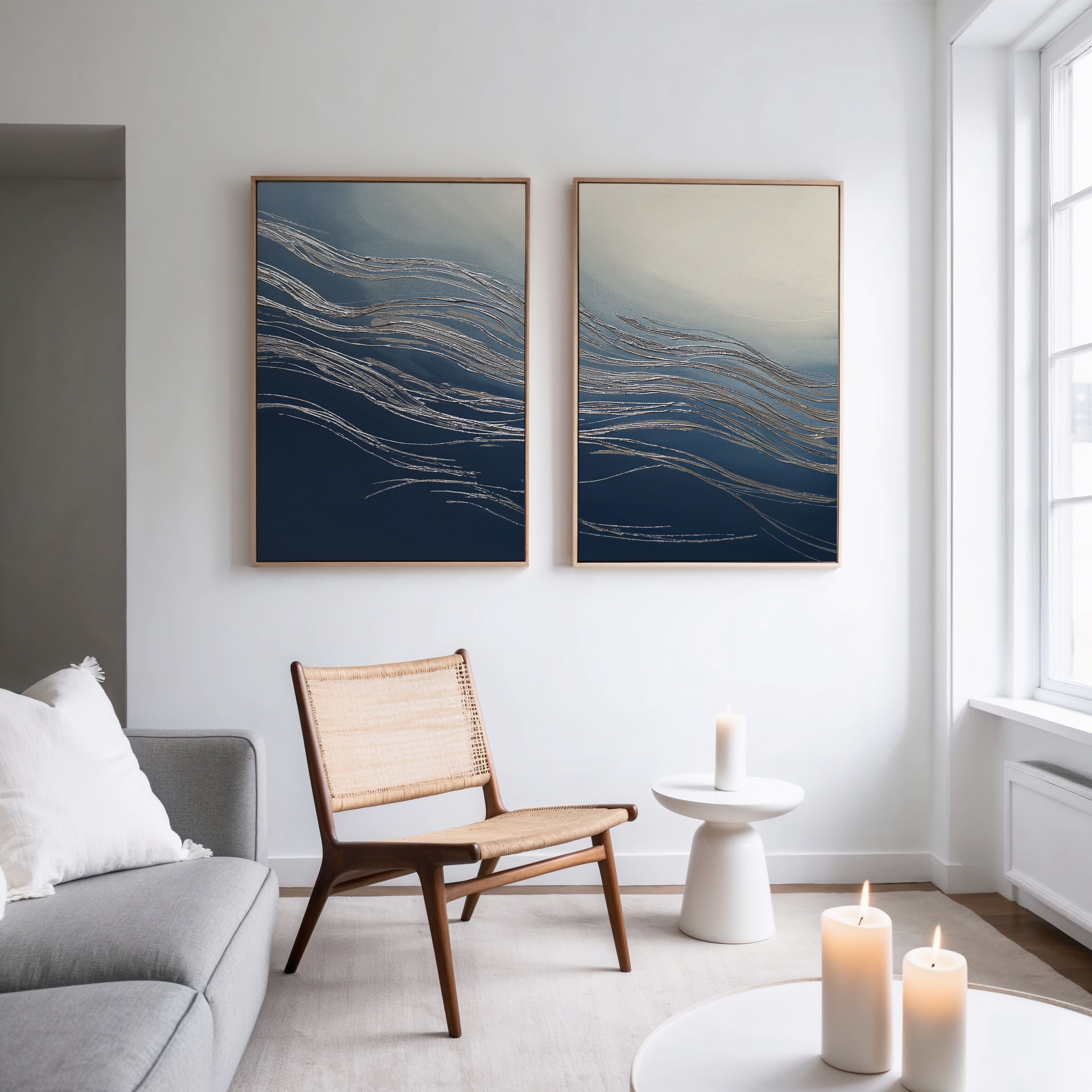A modern living room with a gray sofa, woven chair, and small white side table with a lit candle exudes contemporary style. Natural light streams in from large windows, illuminating the Silver Current - Set Of 2 wall art above.