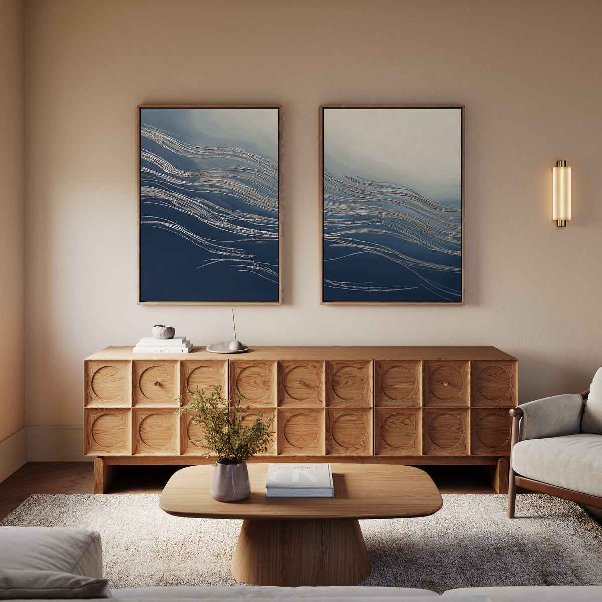 A modern living room with contemporary sophistication features a wooden sideboard, low coffee table, neutral-toned sofa and armchair, abstract blue wall art, a wall sconce light, and the Silver Current - Set Of 2; greenery in a vase decorates the table.