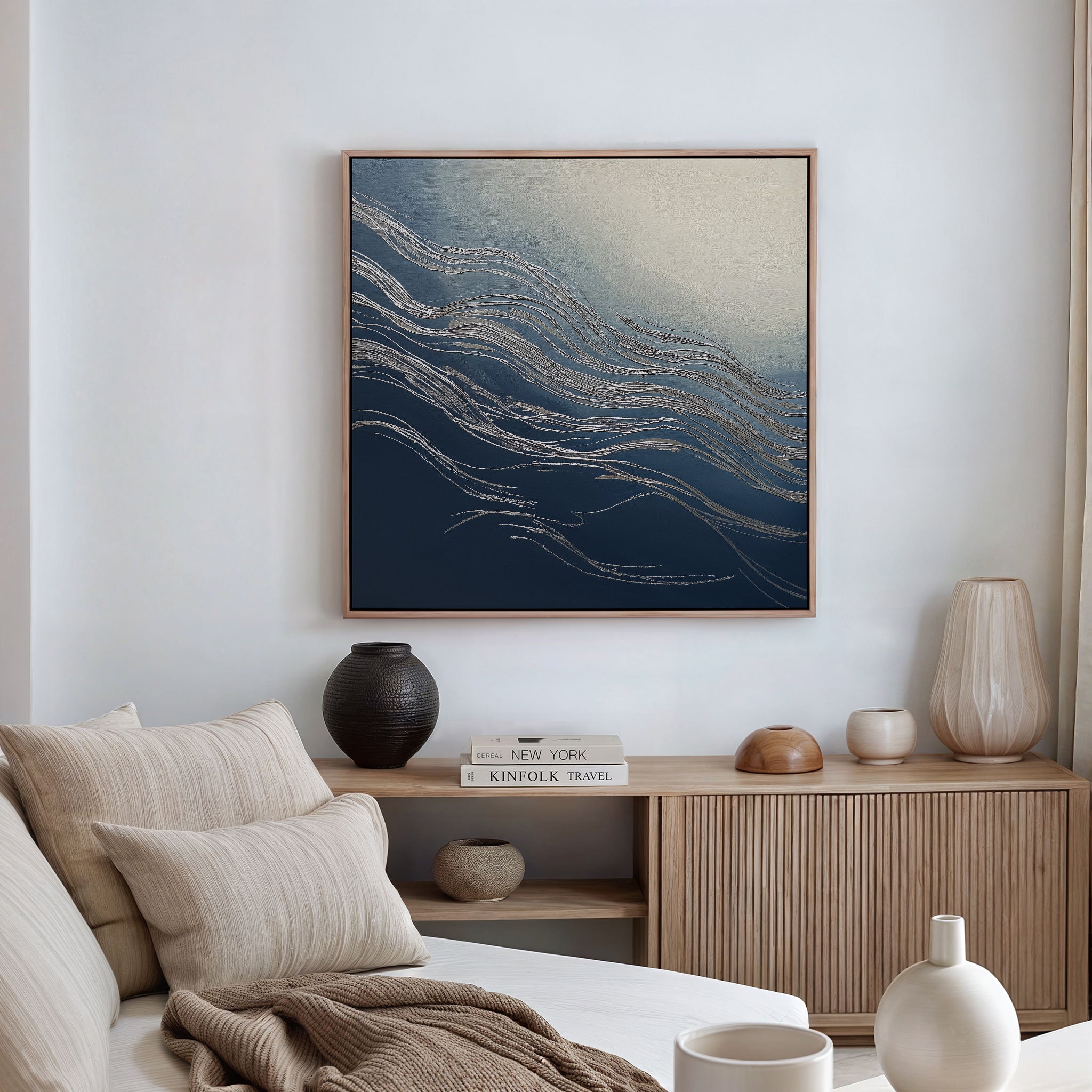 A modern living room with neutral furniture, a wooden sideboard, and decorative ceramics is enhanced by the Silver Current Canvas Art - Square, featuring wavy silver lines on blue and cream tones for a sophisticated contemporary touch.