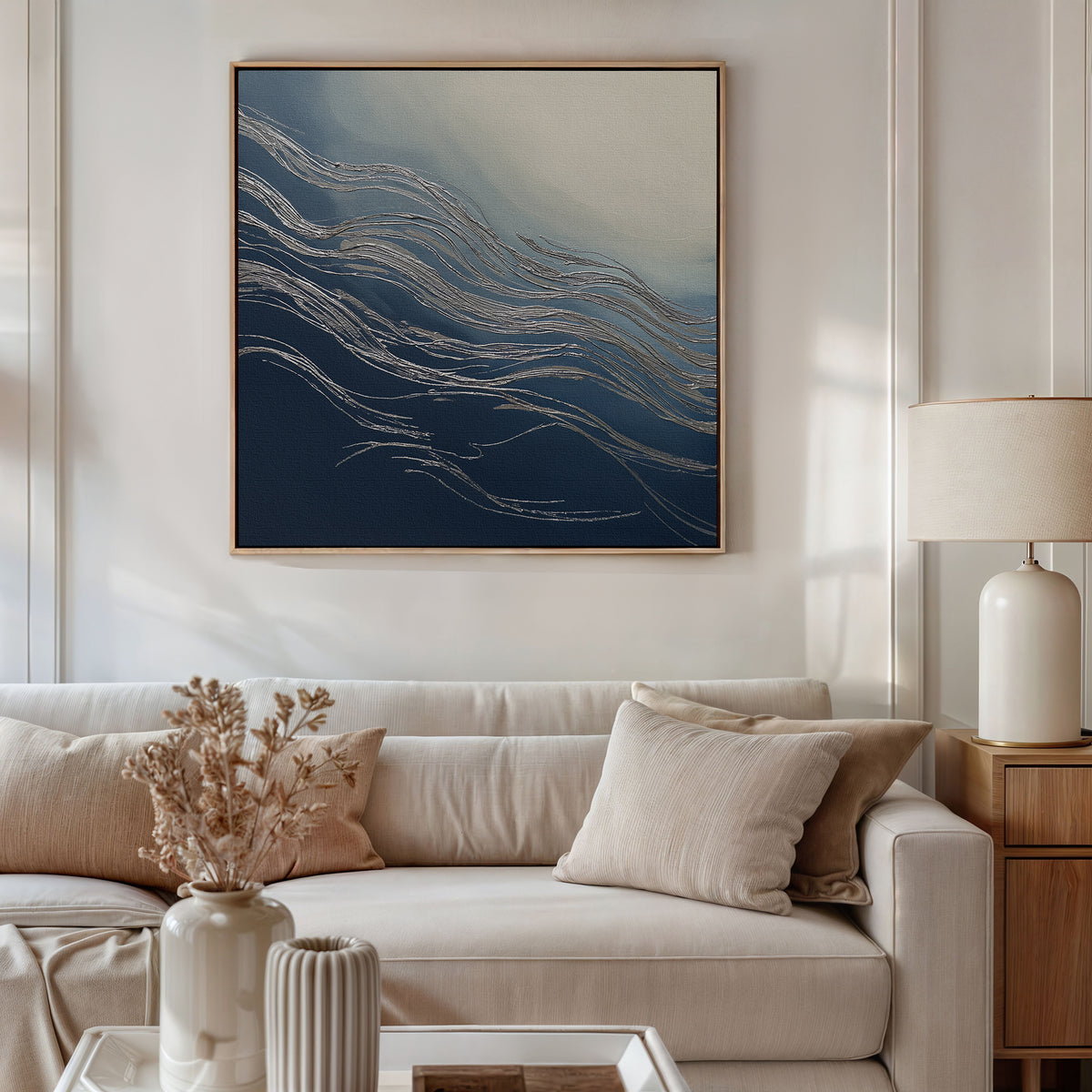 A cozy living room with a beige sofa, neutral pillows, a side table with a cream lamp, dried flowers in vases, and the Silver Current Canvas Art - Square featuring flowing blue and white lines brings contemporary sophistication.