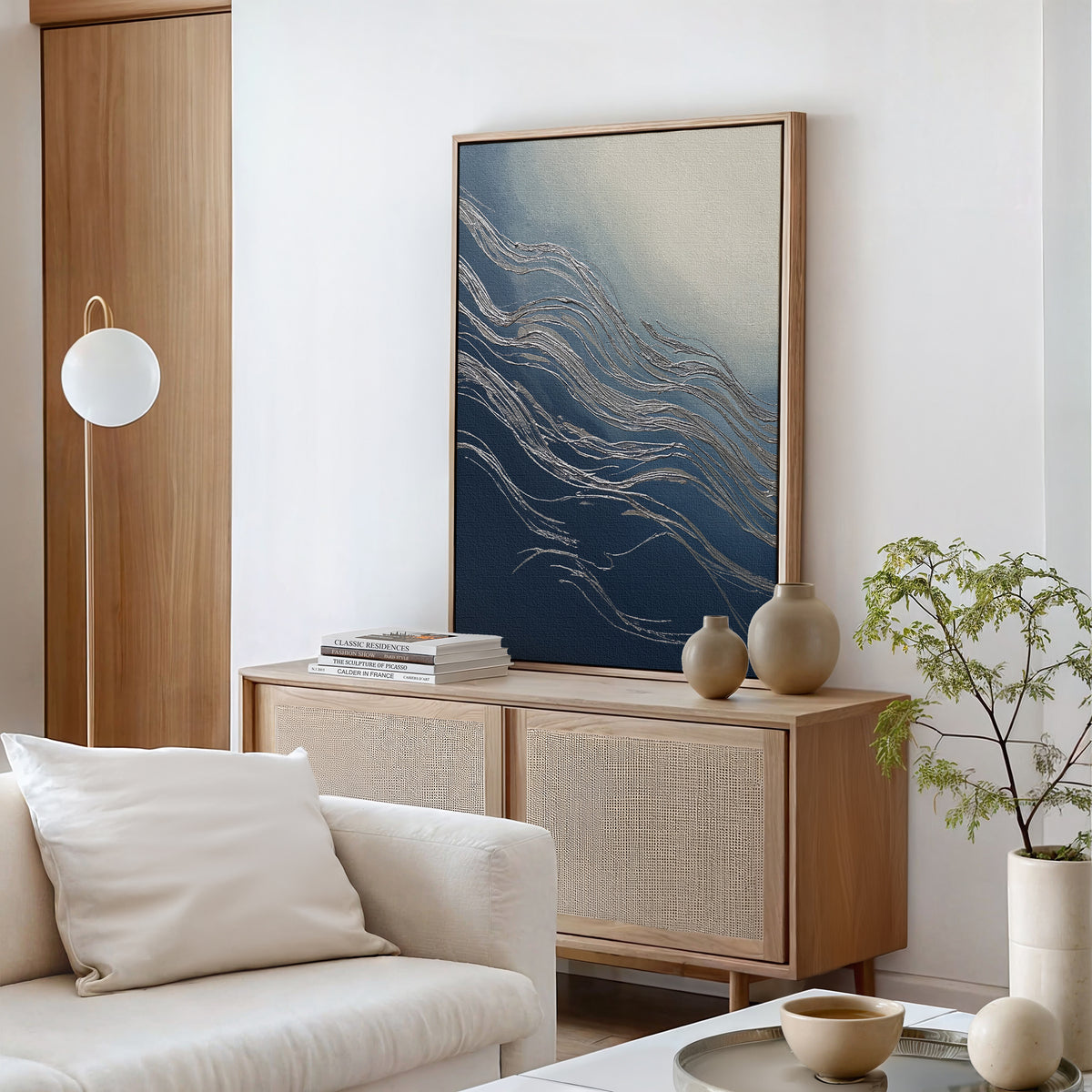 A modern living room featuring the Silver Current Canvas Art - Vertical, a light wood sideboard, neutral vases, books, a white sofa with cushion, a round hanging lamp, and a small plant on a round table for contemporary sophistication.