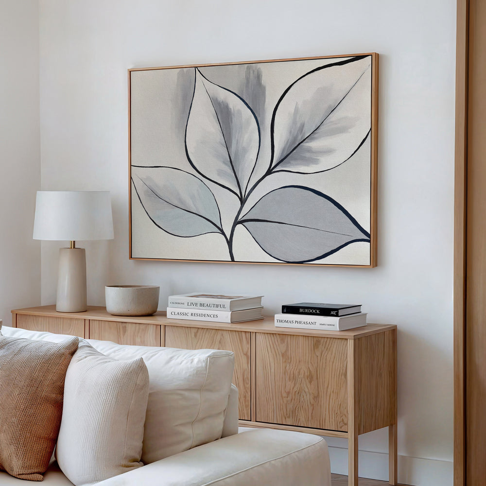 A minimalist living room with a light wood sideboard, neutral decor, stacked books, a white lamp, and the Silver Leaves Canvas Art - Horizontal in shades of gray and black—perfect for modern home decor on a white wall.