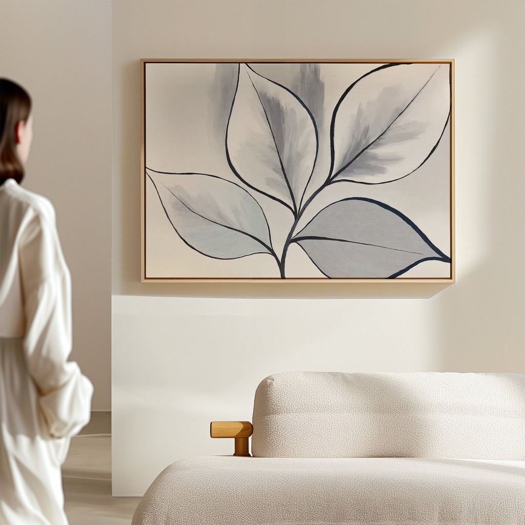 A person in a white shirt stands near a light sofa, admiring the Silver Leaves Canvas Art - Horizontal—a large botanical wall decor piece with abstract black-outlined leaves, perfect for modern, minimalist home interiors.