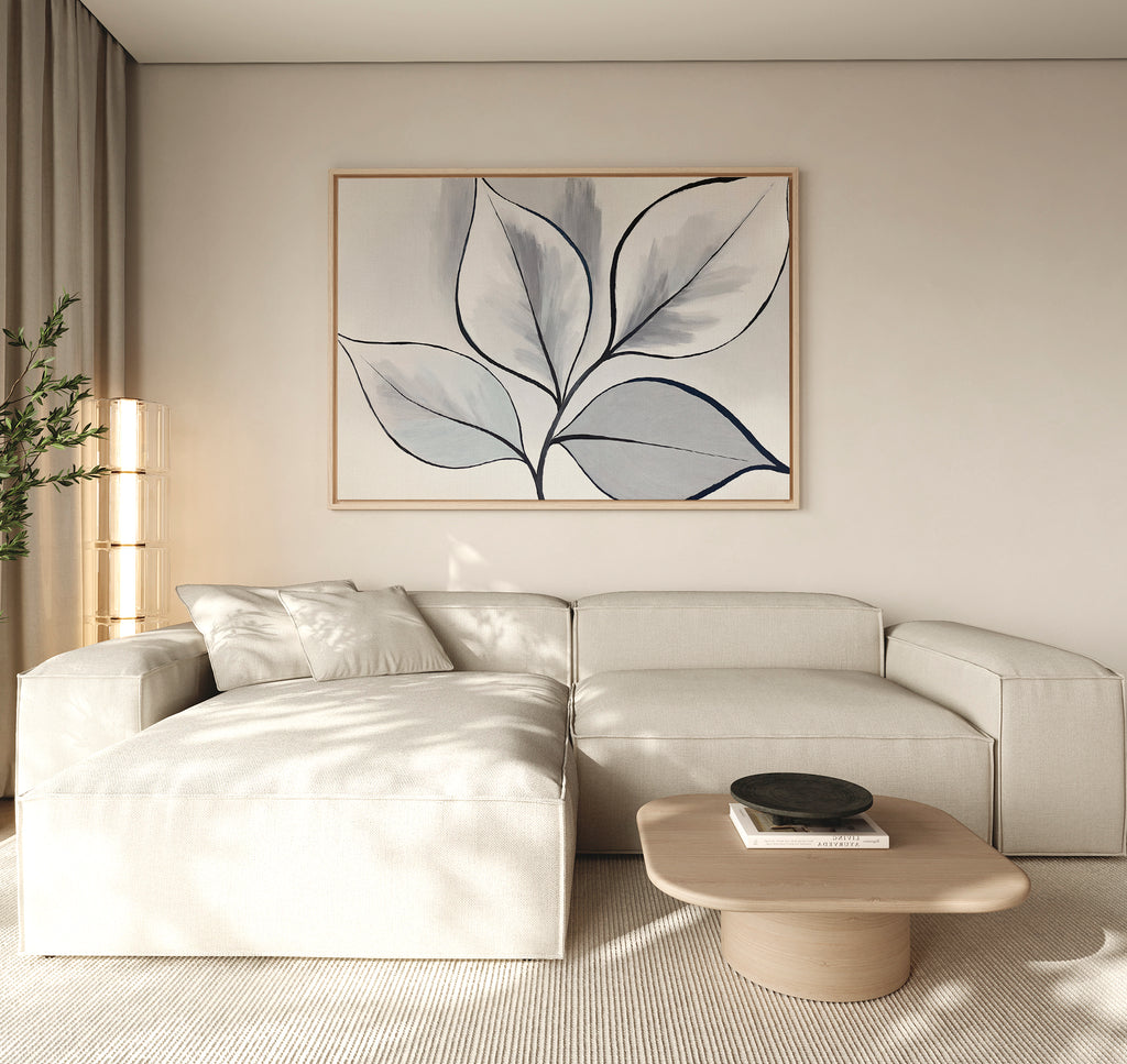A modern living room features a light beige sectional, wooden coffee table with black bowl, neutral rug, tall plant, and the Silver Leaves Canvas Art - Horizontal on the cream wall. Perfect for modern decor lovers as sunlight softly filters in.