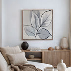 A modern living room with neutral tones, featuring the Silver Leaves Canvas Art - Square as minimalist wall décor, a wooden sideboard with books and ceramic vases, plus a beige sofa with textured cushions for stylish modern home decor.