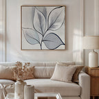 A modern living room with a white sofa, beige cushions, and a lamp on a wooden side table. Above the sofa, the Silver Leaves Canvas Art - Square adds a touch of minimalist decor in neutral tones for a contemporary finish.