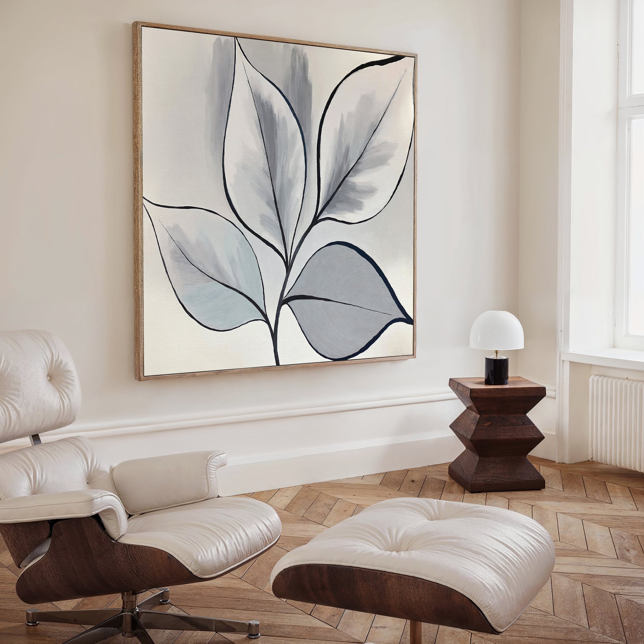 A modern living room featuring Silver Leaves Canvas Art - Square, a white leather lounge chair with ottoman, a geometric wooden side table, and a small lamp by a bright window.