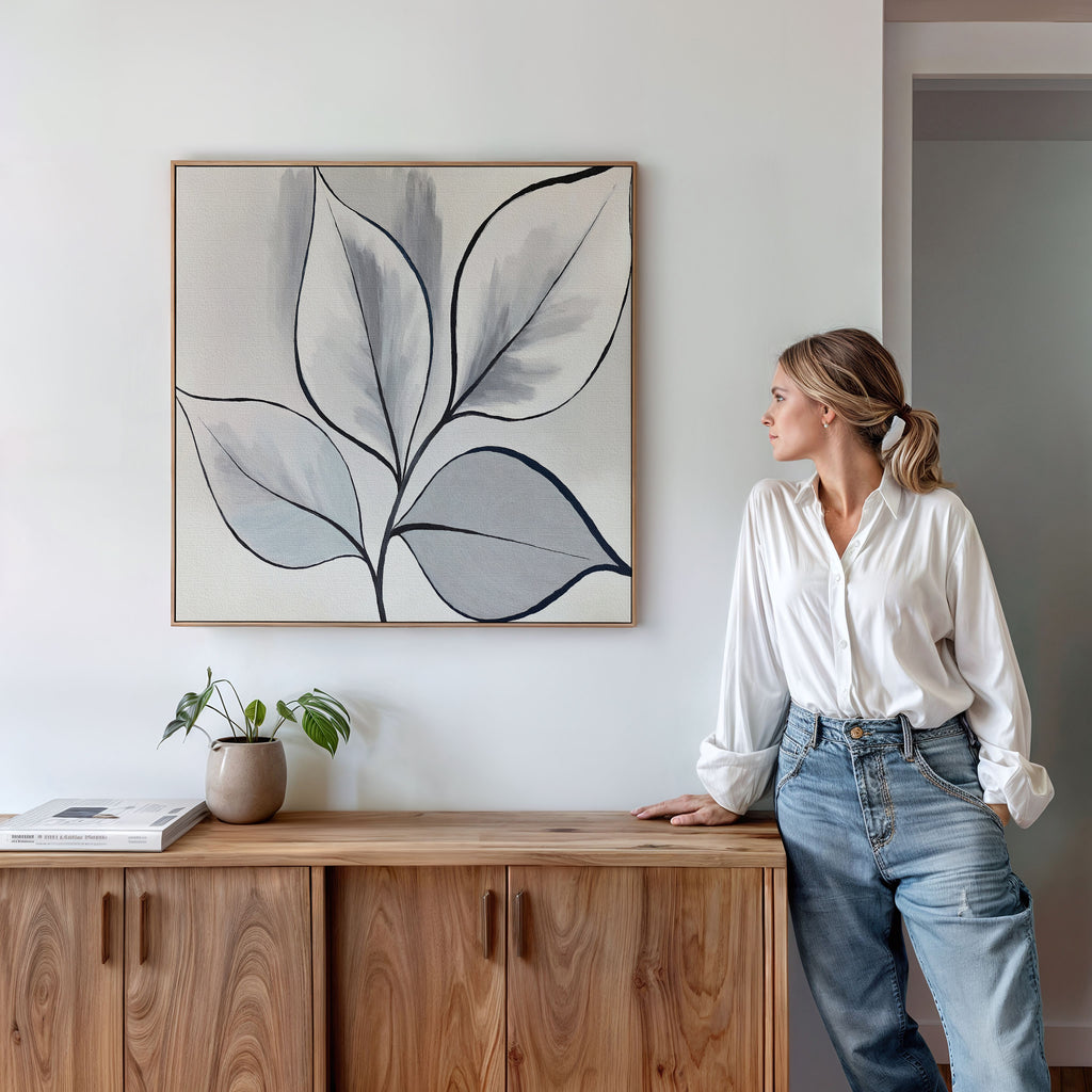 A woman in a white blouse and jeans leans against a wooden cabinet, admiring the Silver Leaves Canvas Art - Square. The minimalist wall art, paired with a small potted plant, elevates the modern decor of her living space.