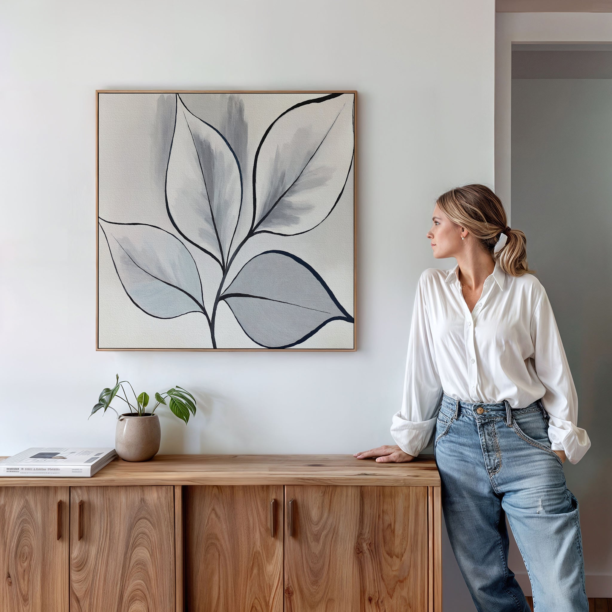 A woman in a white blouse and jeans leans against a wooden cabinet, admiring the Silver Leaves Canvas Art - Square. The minimalist wall art, paired with a small potted plant, elevates the modern decor of her living space.