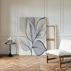 A minimalist room with herringbone wood floors, white paneled walls, a modern white chair, black round side table with candles, and the Silver Leaves Canvas Art - Vertical in muted tones leaning against the wall as decor.