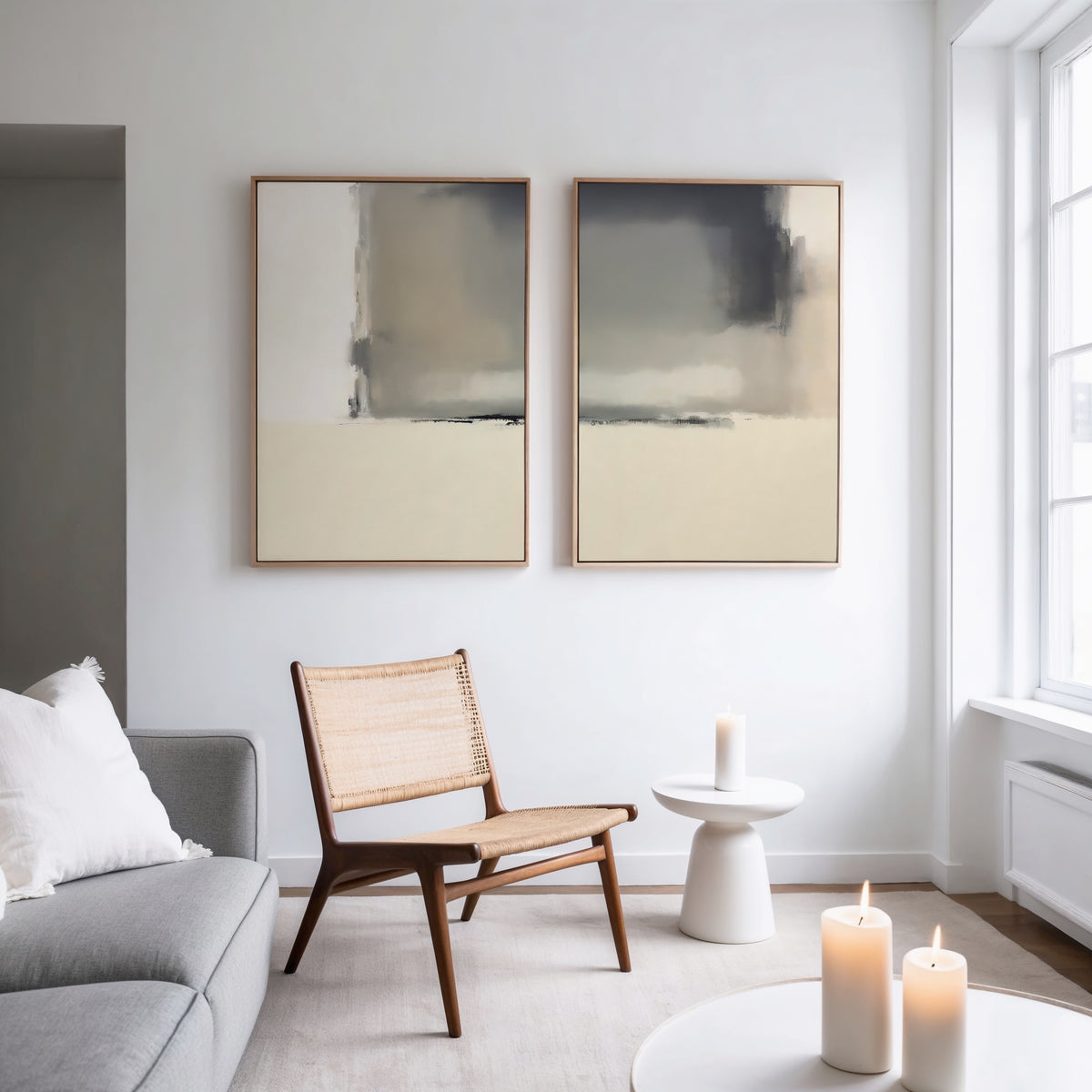A minimal living room features a woven chair, round white side table with a lit candle, and the Smoke Boundary - Set Of 2 abstract wall art. A gray sofa with white cushions and more candles are seen as natural light streams in.