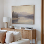A minimalist living room with a wooden sideboard, beige sofa, table lamp, stacked books, and the Smoky Peaks Canvas Art—a vintage-inspired landscape print of a misty lake with trees and mountains—displayed on the wall.