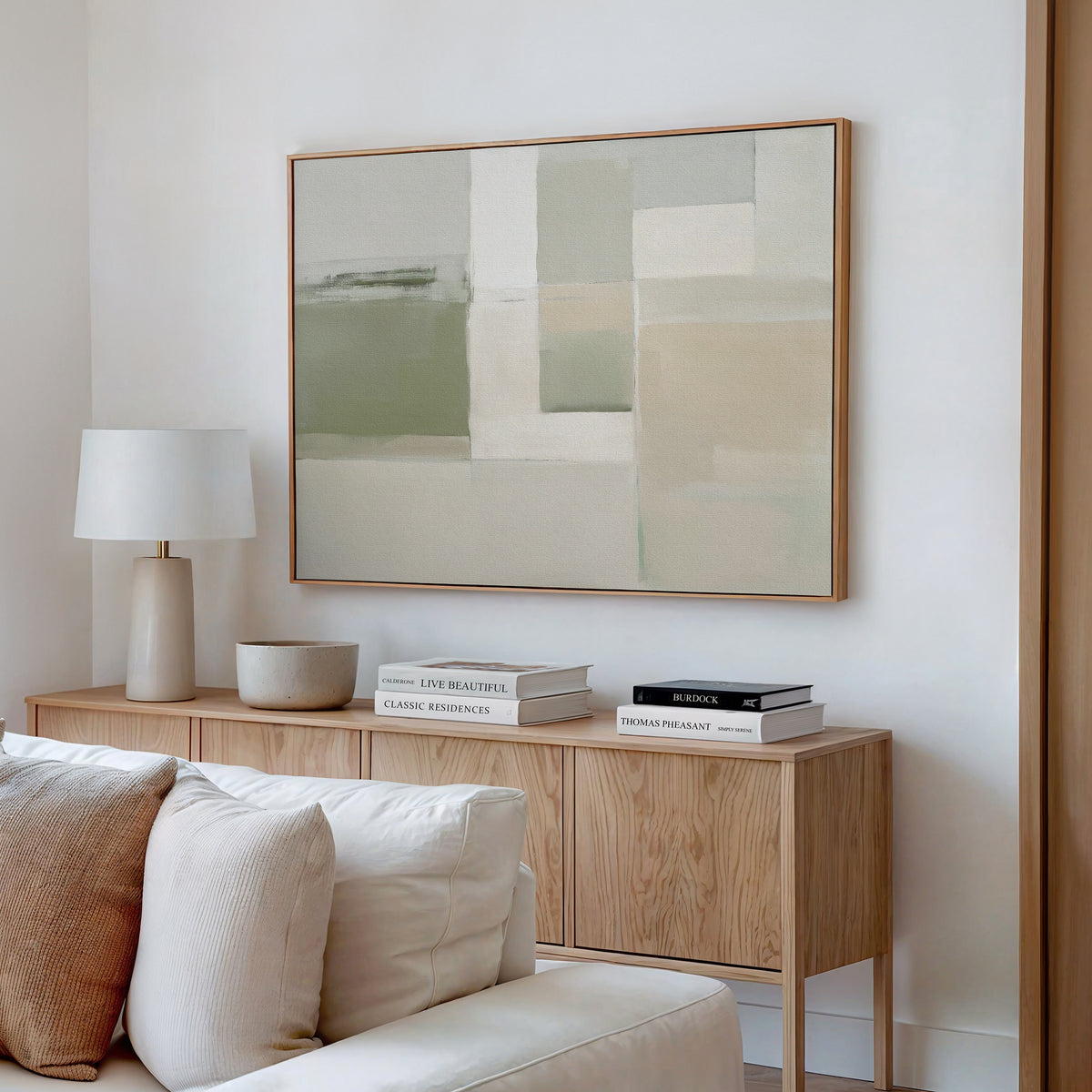 A minimalist living room features a light wood sideboard with books, a white lamp, and a bowl. Above, the Soft Geometric Planes Canvas Art - Horizontal hangs on a white wall. A cream sofa sits in the foreground.