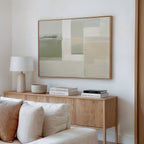 A minimalist living room features a light wood sideboard with books, a white lamp, and a bowl. Above, the Soft Geometric Planes Canvas Art - Horizontal hangs on a white wall. A cream sofa sits in the foreground.