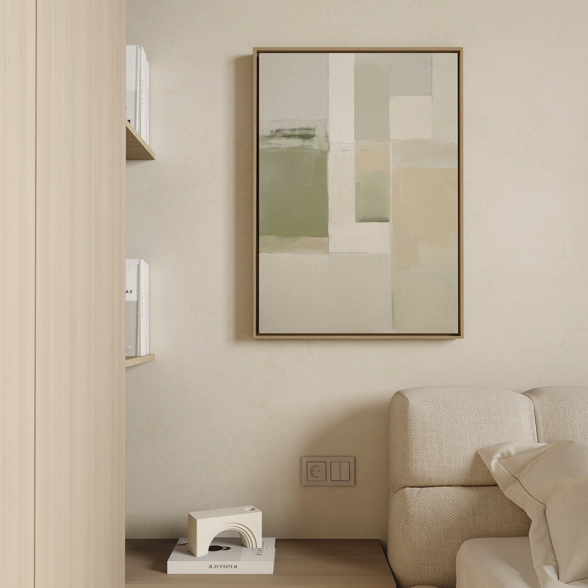 Minimalist living space with a beige wall, contemporary style, part of a light sofa, books on a small shelf, and the Soft Geometric Planes Canvas Art - Vertical featuring abstract green and cream tones as the focal point.