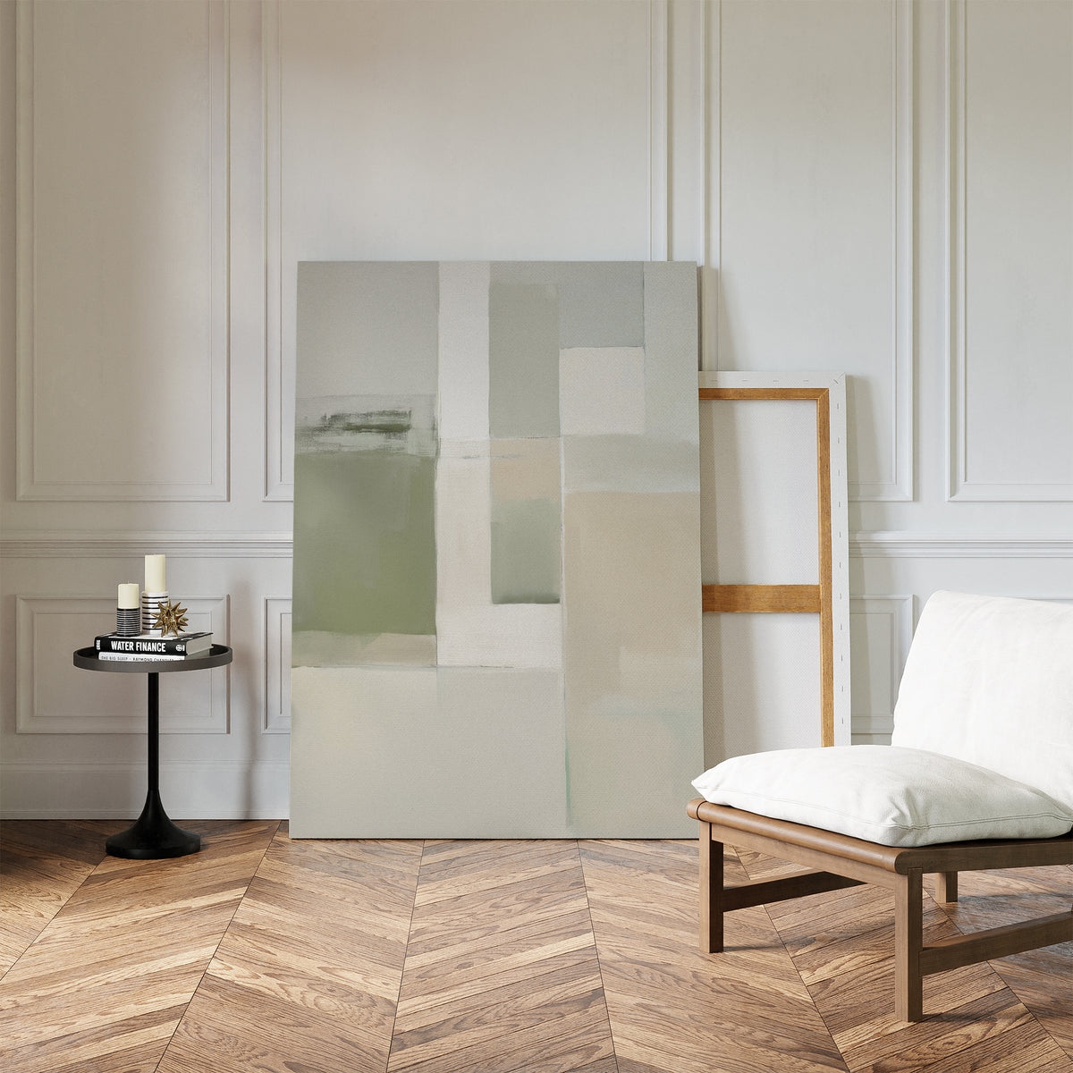 A minimalist room features a white cushioned chair, a small round black side table with candles and books, and the Soft Geometric Planes Canvas Art - Vertical in sage green, leaning against a white paneled wall on herringbone wood flooring.