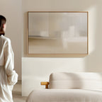A person in a white shirt stands in a bright, minimal room with a beige sofa and the Soft Layers Canvas Art - Horizontal, featuring neutral tones, displayed on the wall.