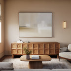 A modern living room with a neutral palette features a wooden sideboard with circular patterns, the Soft Layers Canvas Art - Horizontal above it, a coffee table decorated with flowers, a sofa, and a soft wall-mounted light fixture.