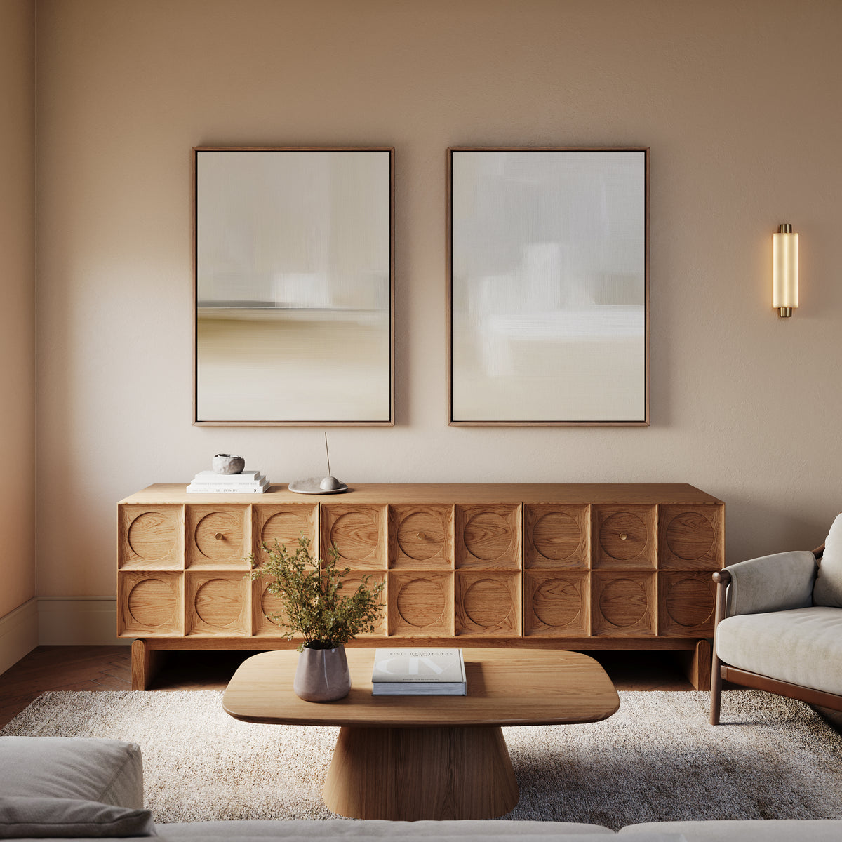 A modern living room features a wooden coffee table, textured sideboard, light armchair, and Soft Layers - Set Of 2 minimalist abstract prints on the wall, accented by a vase of greenery and soft, warm lighting.