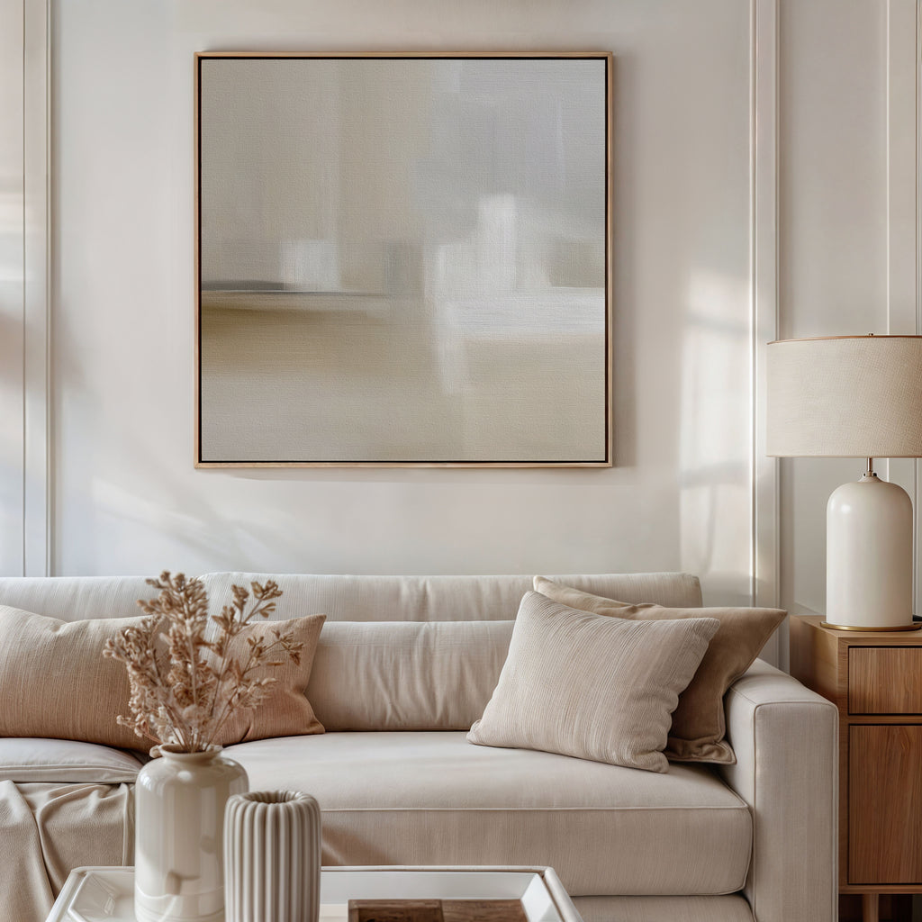 A bright, neutral-toned living room with a beige sofa, cream pillows, and a light wood side table with a white lamp. On the wall hangs the Soft Layers Canvas Art - Square above a coffee table with a vase of dried flowers.