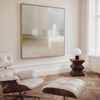 A modern, minimalist room with a cream leather lounge chair and ottoman, geometric wooden side table, small white lamp by a large window, and the Soft Layers Canvas Art - Square featuring a neutral abstract print above.