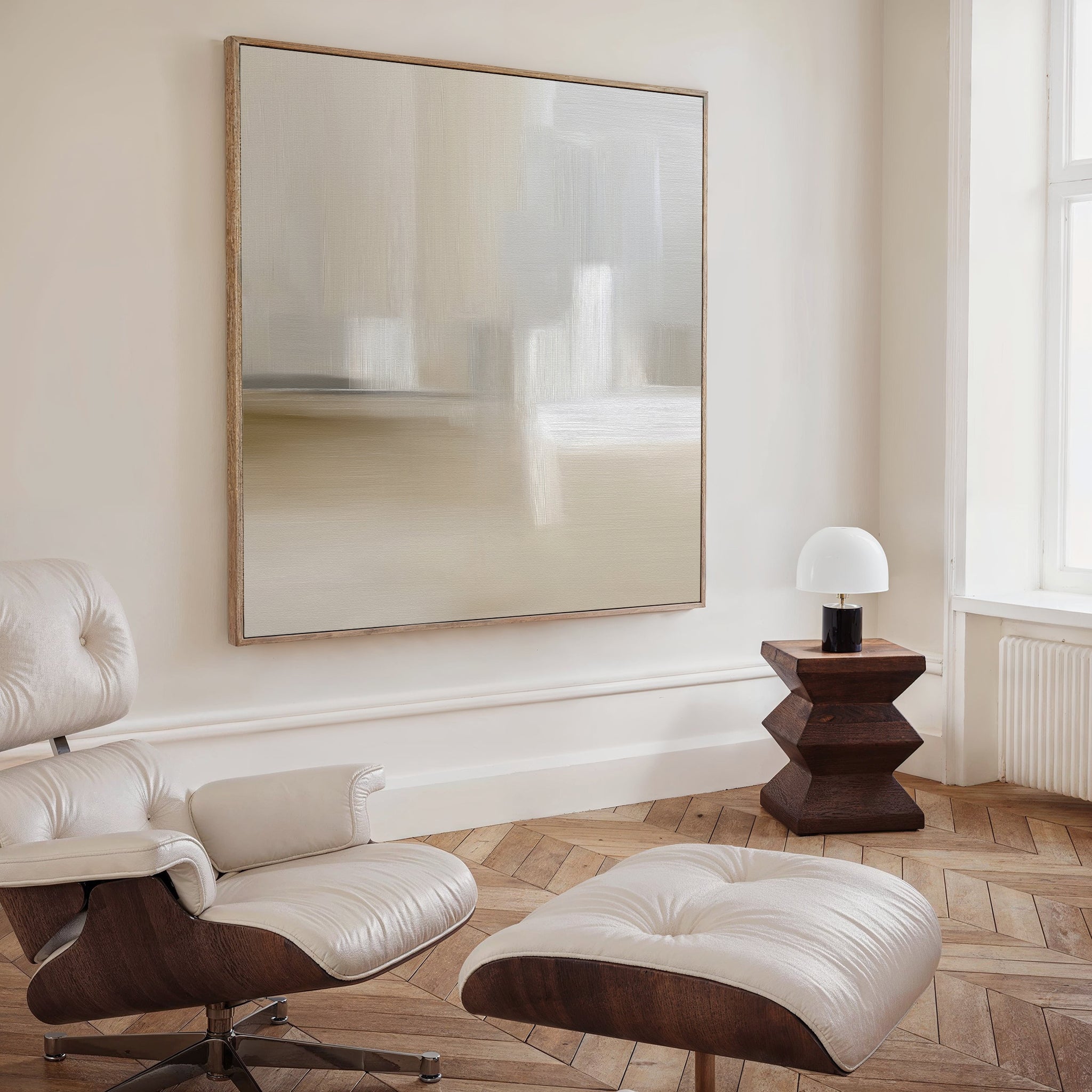A modern, minimalist room with a cream leather lounge chair and ottoman, geometric wooden side table, small white lamp by a large window, and the Soft Layers Canvas Art - Square featuring a neutral abstract print above.
