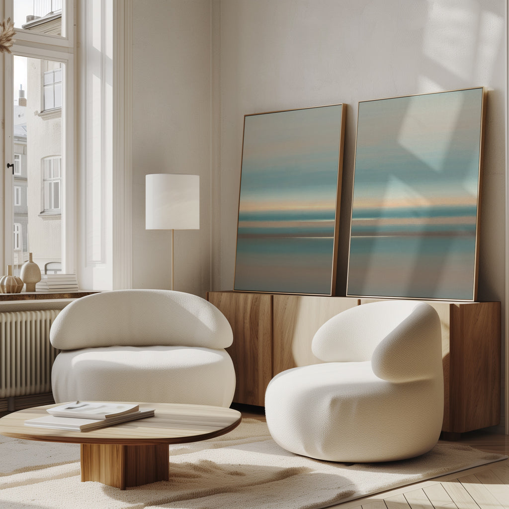 A modern living room with two curved white armchairs, a round wooden coffee table, and Soft Tide Layers - Set Of 2 abstract prints in blue and beige on a wooden cabinet by a tall window. Sunlight fills this serene Nordic Park-inspired space.