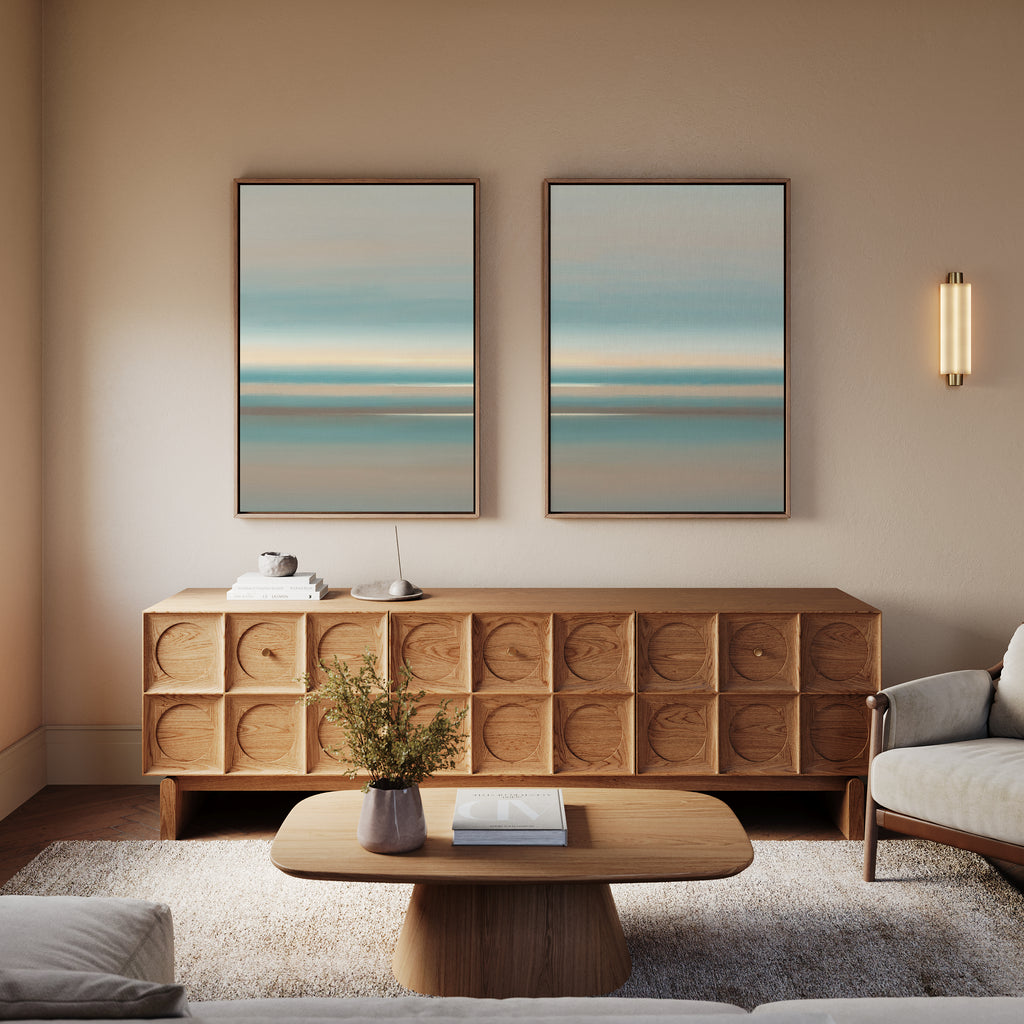 A modern living room with a wooden sideboard, Soft Tide Layers - Set Of 2 wall art in blue and beige above it, a round wooden coffee table with books and a vase, and a light armchair on a rug. Warm lighting adds coziness.