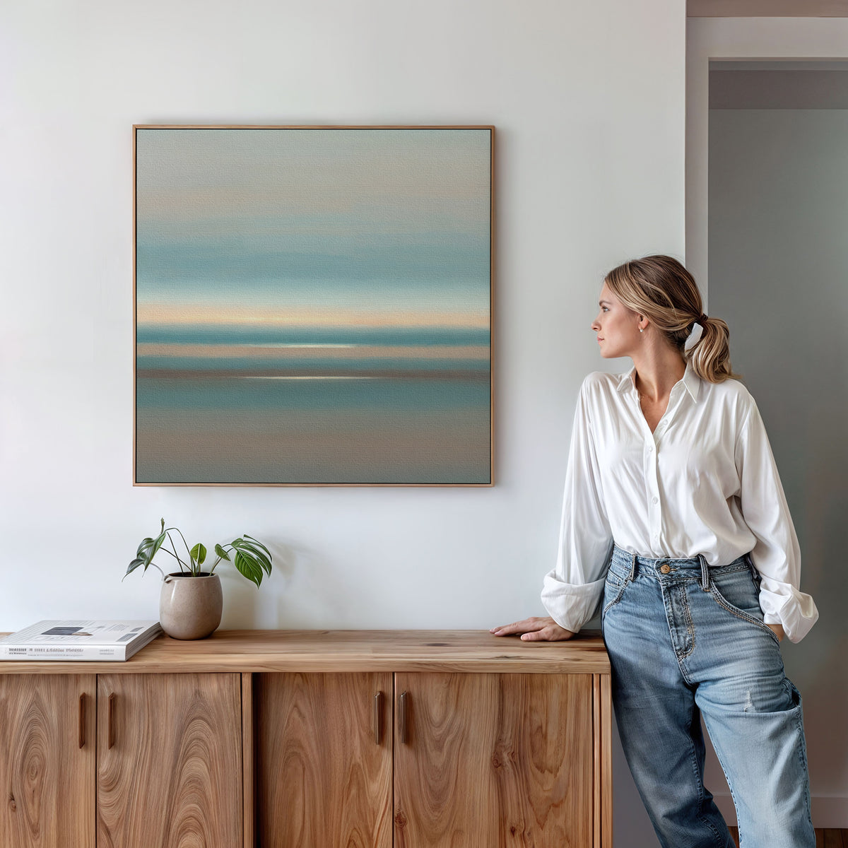 A woman in a white blouse and jeans stands by a wooden sideboard, admiring the Soft Tide Layers Canvas Art - Square, with calming blue and beige tones, coastal wall art on a white wall. A small plant and books rest on the sideboard.